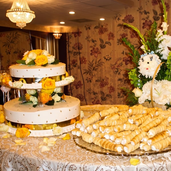A two-tier white cake with yellow roses and a platter of cream horns on a decorated table.