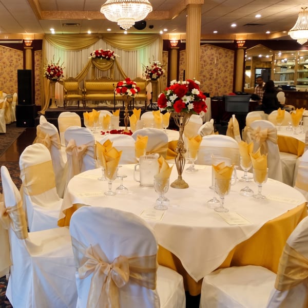 Elegant banquet hall with white and gold tables, chairs, and a decorated stage with red floral arrangements.