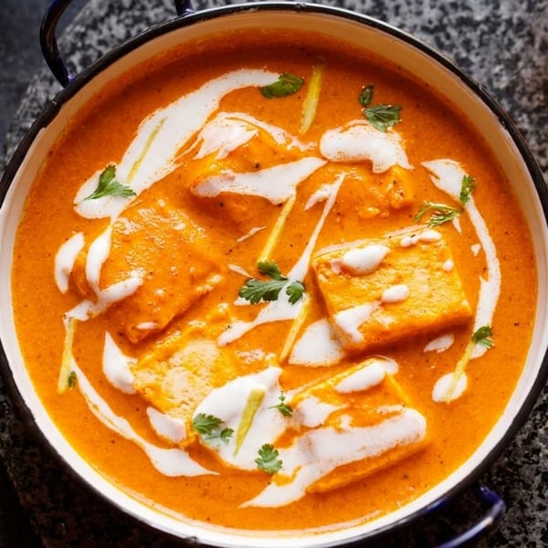 Overhead view of paneer butter masala in a white bowl with black rim, garnished with cream and herbs.